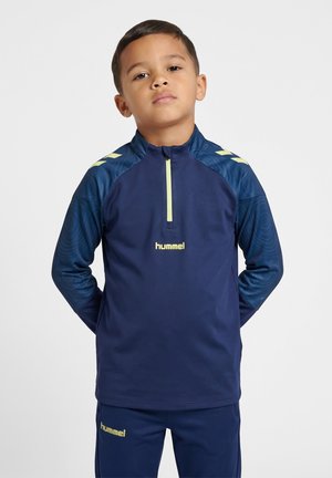 Ung dreng iført en marineblå hummel sportjakke og bukser med lysegule accenter, stående med hænderne bag ryggen.