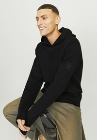 Sweatshirt à capuche noire avec des manches côtelées et un détail en patch sur le bras, associé à un pantalon beige texturé.