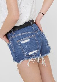 Shorts de mezclilla con un dobladillo deshilachado, parches desgastados y bolsillos traseros. Incluye un cinturón negro y una blusa de color claro con un patrón decorativo.