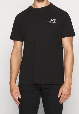 Man wearing a black short-sleeve t-shirt with white EA7 Emporio Armani logo on the chest and black pants.