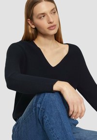 Schwarzer Strickpullover mit V-Ausschnitt, langen Ärmeln und gerippten Bündchen, kombiniert mit blauen Jeans. Schlicht, strukturiert.