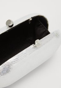 Mała, srebrna torebka kopertówka o teksturowanej powierzchni z czarną podszewką i okrągłym, srebrnym zapięciem, pokazana otwarta na białym tle.