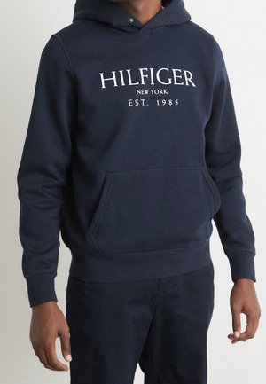 Osoba nosząca granatową bluzę z kapturem z napisem "Hilfiger New York Est. 1985" nadrukowanym na białym tle na piersi, połączoną z pasującymi granatowymi spodniami.