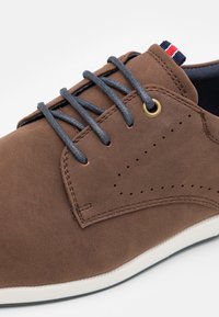 Zapato de ante marrón con cordones grises y un ojal dorado, con detalles perforados y una suela de goma blanca. Acentuado con una etiqueta roja y azul.