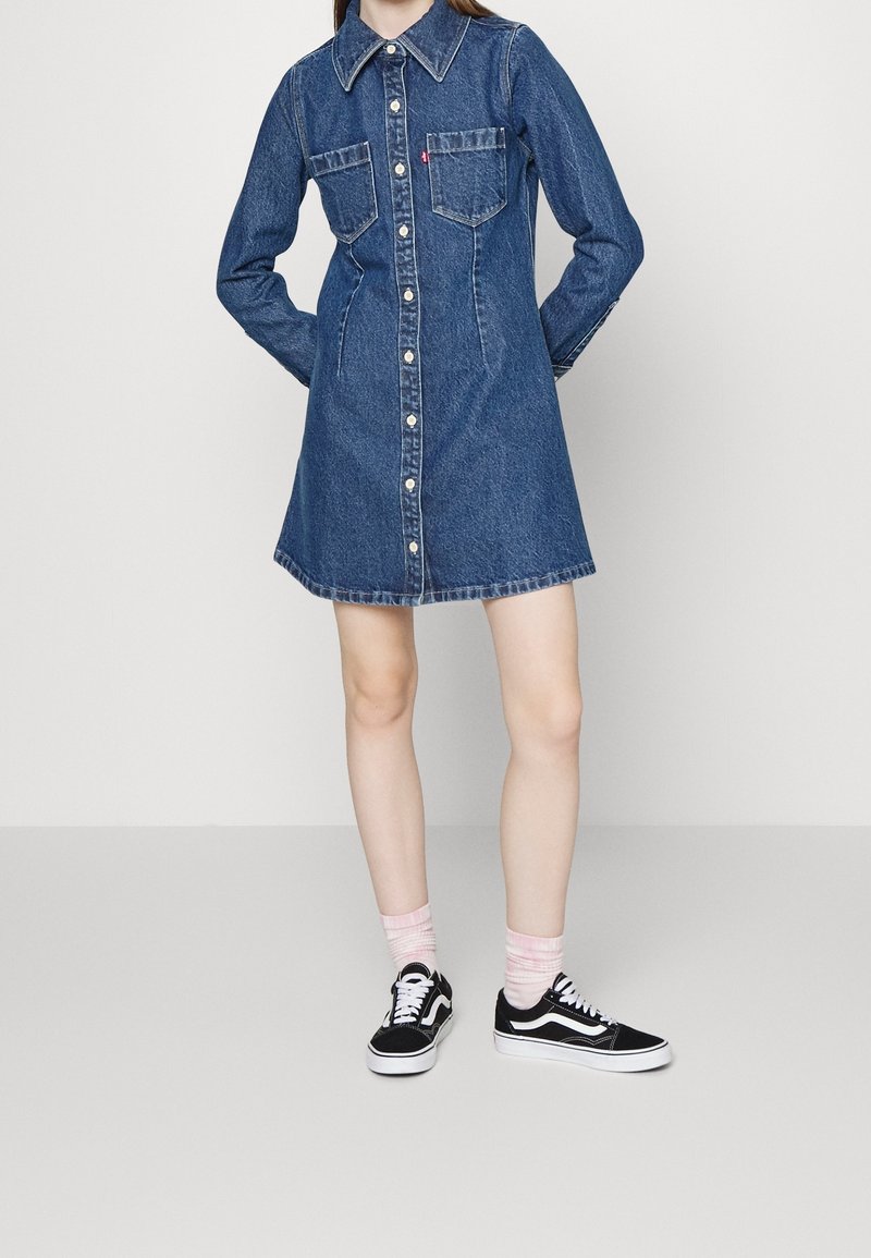 Robe chemise en denim bleu foncé, avec un devant boutonné, deux poches poitrine, et une coupe décontractée. Associée à des baskets noires et des chaussettes roses.