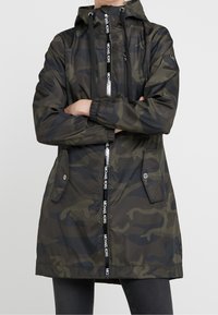 Vrouw in een donkergroen en zwart camouflage-hoodie met voorzakken en een ritssluiting, gelabeld "Michael Kors."
