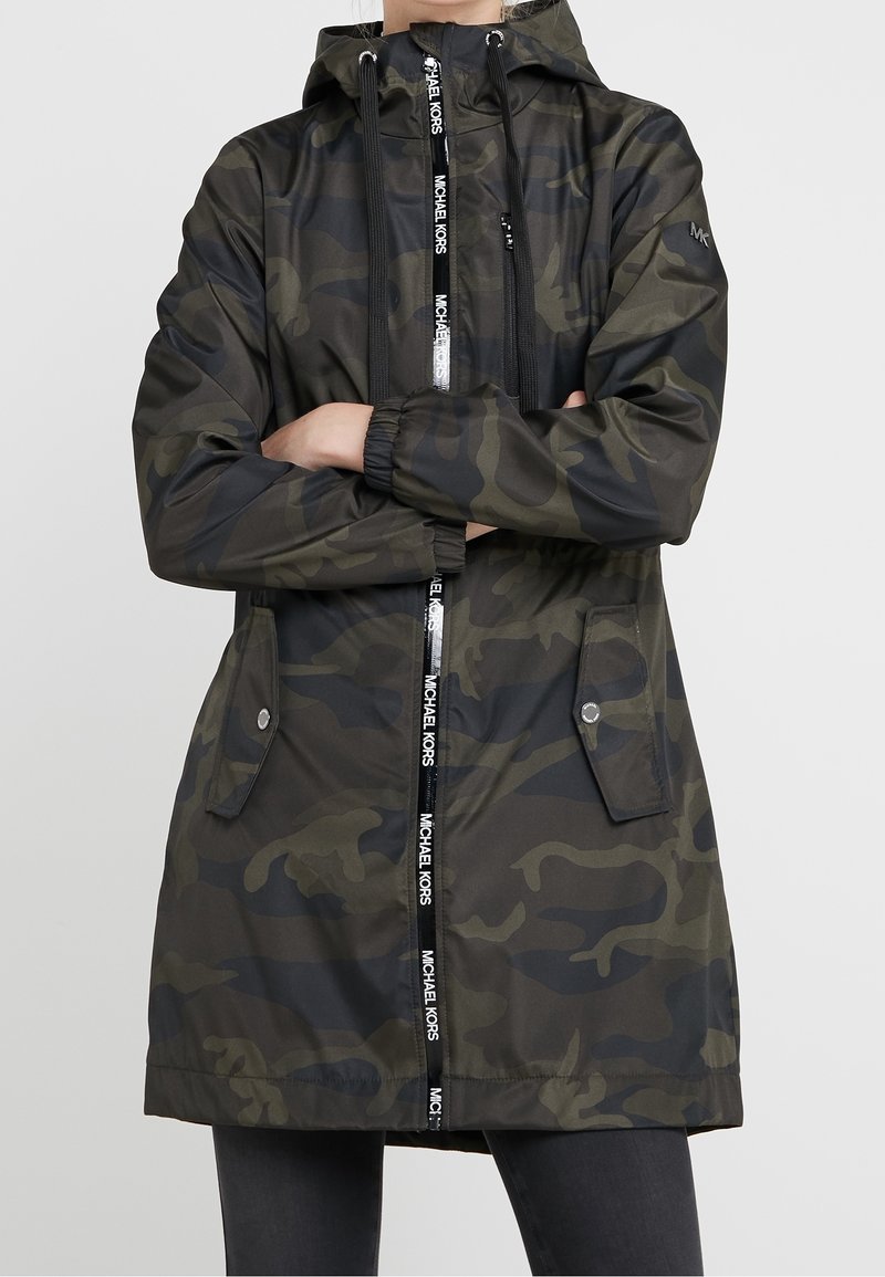 Vrouw in een donkergroen en zwart camouflage-hoodie met voorzakken en een ritssluiting, gelabeld "Michael Kors."