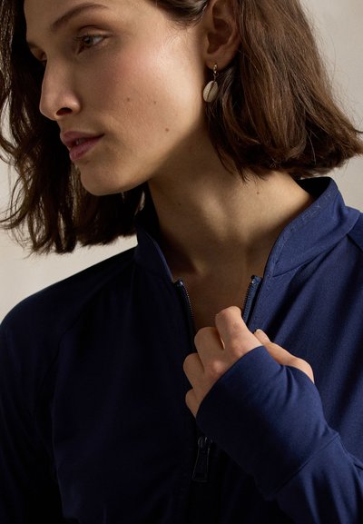 Κοντινό πλάνο μιας γυναίκας με κοντά καστανά μαλλιά που φοράει μπλε μπλουζάκι με φερμουάρ και κρατά το φερμουάρ κοντά στον λαιμό της, δείχνοντας ένα σκουλαρίκι σε σχήμα κοχυλιού.