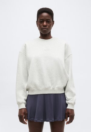 Νεαρό άτομο μαύρης καταγωγής που φοράει ανοιχτό γκρι φαρδύ φούτερ και κοντή σκουρόχρωμη πλισέ φούστα, στέκεται μπροστά σε απλό φόντο.