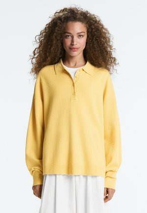 Žena s kučeravými vlasmi, ktorá nosí voľný žltý polo sveter s dlhým rukávom cez biele šaty, stojaca pred jednofarebným svetlým pozadím.