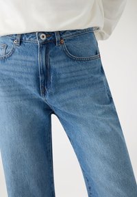 Jeans in denim azzurro chiaro con vestibilità classica, vita alta, dettagli in rame e tessuto leggermente scolorito. Tasche frontali visibili e chiusura con bottone.