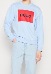 Sweat-shirt bleu clair avec un graphisme rectangulaire rouge affichant le mot "HUGO" en lettres noires épaisses. Poignets et ourlet côtelés.