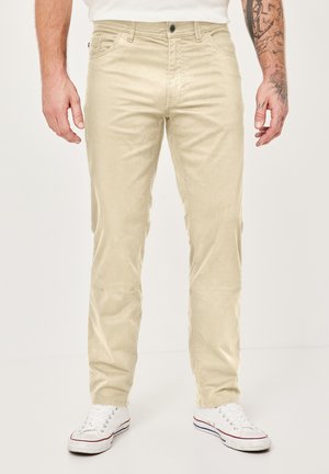 Mann trägt hellbeige Freizeithose und weiße Sneakers, steht vor einem schlichten hellen Hintergrund, mit sichtbaren Tattoos am rechten Unterarm.