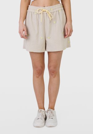 Femme portant un short beige avec cordon de serrage, orné de petits motifs de citrons jaunes, et des baskets blanches, debout devant un fond uni.