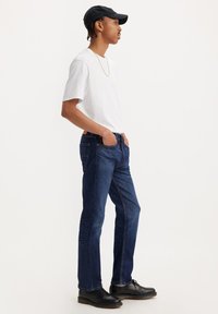Camiseta blanca de manga corta, jeans de mezclilla azul oscuro, gorra negra y zapatos negros. Diseño de pierna recta con bolsillos y una marca mínima.