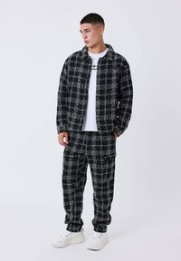 Jonge man in zwart-witte geblokte jas en broek met witte sneakers en een grafisch wit T-shirt, staande tegen een effen achtergrond.