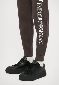 Juodi odiniai sportbačiai su storesniais padais, derinami su rudos spalvos sportinėmis kelnėmis, kuriose yra baltas "Emporio Armani" logotipas ir lygus paviršius.