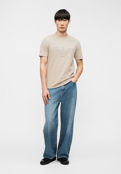 Μπεζ βαμβακερό T-shirt με ανάγλυφο τριγωνικό σχέδιο στο στήθος, συνδυασμένο με μπλε τζιν παντελόνι φαρδιάς γραμμής. Μαύρα παπούτσια ολοκληρώνουν το ντύσιμο.