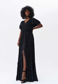 Robe maxi noire avec des manches volantes et un décolleté en V profond. L fente sur le côté dévoile la jambe, avec un détail de taille texturé et un tissu lisse.