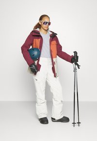 Skikleding met een bordeauxrood, oranje en crèmekleurige jas, witte broek, zwarte handschoenen en skistokken. Een blauwe helm en zonnebril maken de look af.