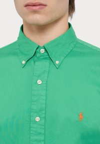 Zelená bavlněná košile s límcem na knoflíky, perleťovými knoflíky a oranžovým vyšitým logem na levé hrudi. Hladká textura.