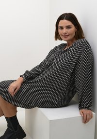 Robe à motifs noir et blanc avec des manches longues, coupe ample, dotée d'un col en V et de formes géométriques subtiles. Portée avec des bottines noires. 