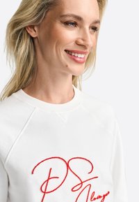 Felpa bianca con testo ricamato rosso. Presenta un collo rotondo e maniche raglan. Tessuto morbido con dettagli di cucitura visibili.
