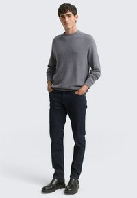 Szary sweter w robótki z okrągłym dekoltem, ciemnoniebieskie spodnie i czarne botki za kostkę. Gładka faktura i luźny krój. Postawa stojąca na jasnym tle.