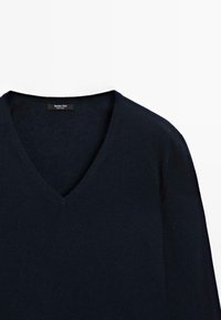 Pull col en V bleu marine en laine douce, présentant une texture lisse et un design minimaliste. Étiquette visible au niveau du col.