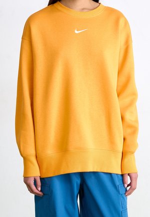 Osoba ubrana w luźną żółtą bluzę Nike z długimi rękawami i niebieskie spodnie typu cargo, stojąca z rękami swobodnie opuszczonymi wzdłuż ciała.