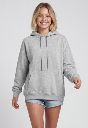 Ung kvinde med blondt hår, smilende, iført en lysegrå hættetrøje og lyseblå denimshorts mod en ensfarvet baggrund.