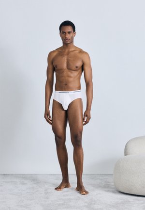 Białe bokserki z czarnym paskiem z logo, o gładkim wzorze, miękkiej tkaninie i dopasowanym kroju, noszone przez męską postać stojącą na jasnym dywanie.