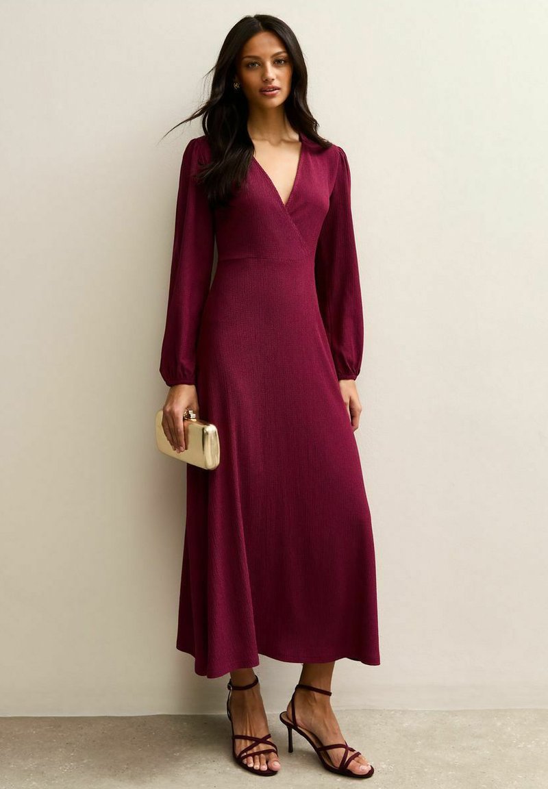 Femme portant une robe midi à manches longues, col en V profond, de couleur rouge foncé, avec des talons à lanières, tenant une petite pochette beige sur un fond uni.