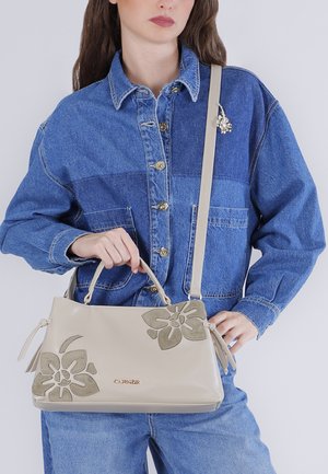 Femme portant une veste en jean patchwork tenant un sac à main beige avec des appliqués floraux et une bandoulière, sur un fond clair.