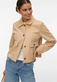 Femme souriante, portant une veste beige boutonnée avec poches à rabat, un haut blanc, un jean bleu et de longues boucles d'oreilles, posant avec une main sur la hanche.