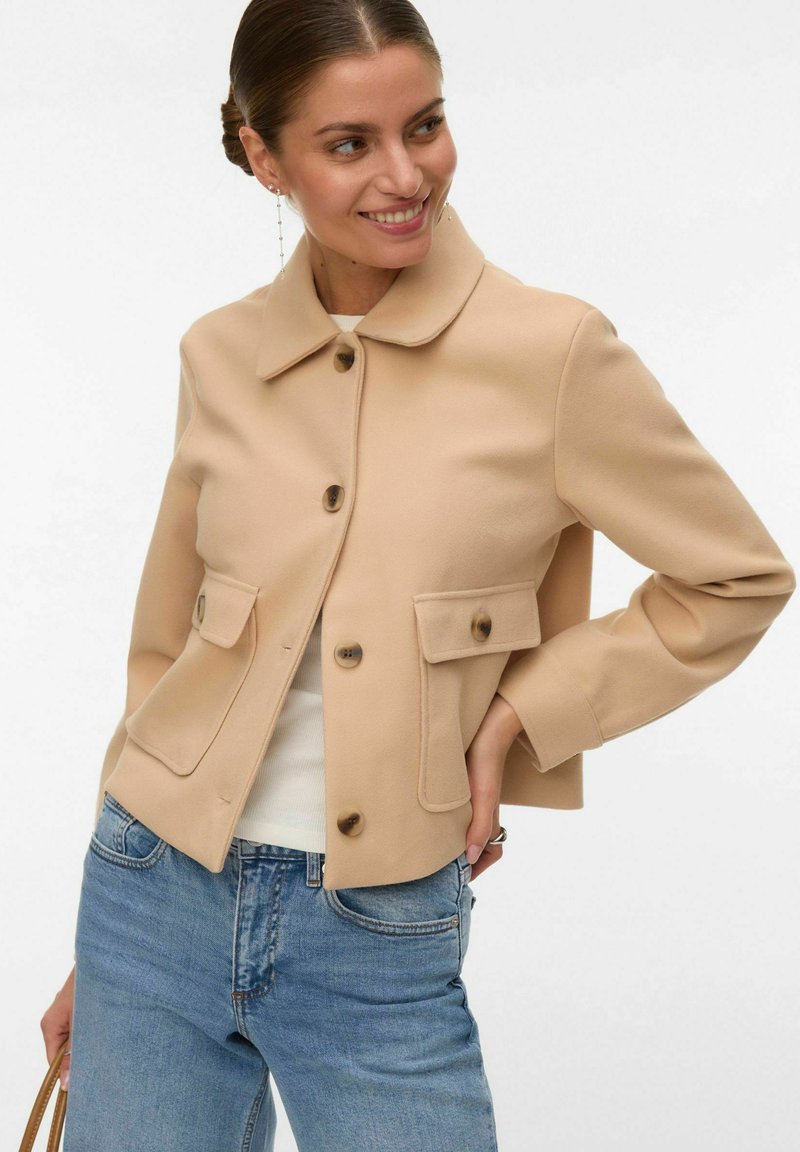 Femme souriante, portant une veste beige boutonnée avec poches à rabat, un haut blanc, un jean bleu et de longues boucles d'oreilles, posant avec une main sur la hanche.