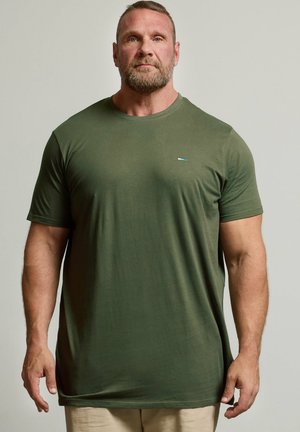 Muskelbepackter Mann mit kurzen Haaren und Bart, der ein einfarbiges olivgrünes T-Shirt und beige Hose trägt und vor einem neutralen Hintergrund steht.