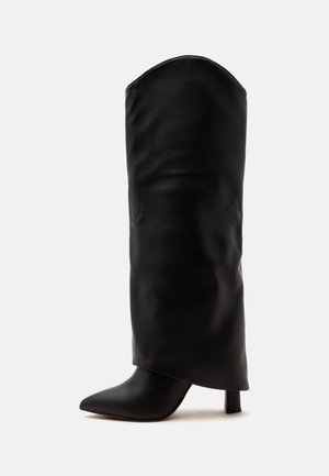 Botte haute en cuir noir avec bout pointu et talon bloc haut sur un fond clair.