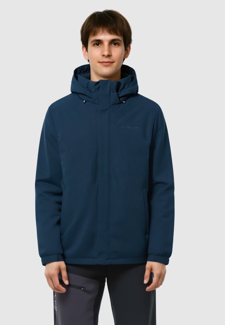 Jeune homme portant une veste à capuche bleu marine et un pantalon sombre, se tenant devant un fond gris clair uni.