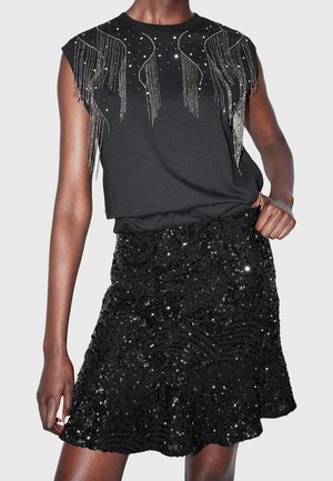 Sort ærmeløs top med sølvperlefringe og sort palietnederdel med blomster- og bølgemønstre, båret af en person med mørk hud.