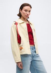 Νεαρή γυναίκα που φοράει κρεμ μπουφάν με κεντημένα σχέδια, κόκκινη υφασμάτινη μπλούζα, ανοιχτόχρωμο μπλε τζιν και μικρό κόκκινο τσαντάκι ώμου.