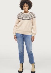 Pull en tricot beige avec un motif géométrique noir et blanc sur l'empiècement, doté de manches longues et d'une coupe décontractée. Assorti à un jean bleu clair.