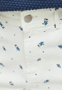 Tissu blanc avec de petits motifs floraux bleus, présentant un bouton et des détails de ceinture d'un pantalon. Pull bleu texturé au-dessus.
