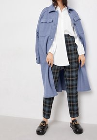 Lichtblauwe wollen jas over een witte blouse, gecombineerd met zwart-blauwe geruite broek en zwarte instapschoenen met een kettingdetail.