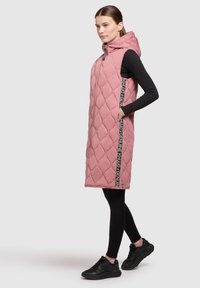 Rosa gesteppte ärmellose Steppweste mit Kapuze, die ein verlängertes Design, seitliche Taschen und einen mit Logo bedruckten Riemen auf der Vorderseite aufweist.