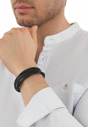 Mann mit leicht stoppeligem Gesicht, der ein weißes Hemd trägt und das Kinn auf die Faust stützt, wodurch ein schwarzes Lederarmband mit mehreren Bändern am Handgelenk sichtbar ist.