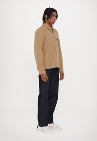 Beige Zip-Jacke mit Kragen und Brusttasche, kombiniert mit dunkelblauen Jeans und weißen Sneakers. Glattes Material mit minimalistischem Design.