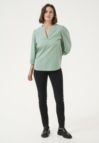 Blouse vert menthe avec un décolleté en V et des manches bouffantes trois-quarts, texturée avec de petits motifs. Associée à un pantalon slim noir et des mocassins noirs.