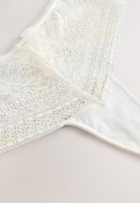 Biancheria intima in pizzo bianco con motivi floreali e bordi smerlati su uno sfondo chiaro, che mette in risalto la delicata trama del tessuto.