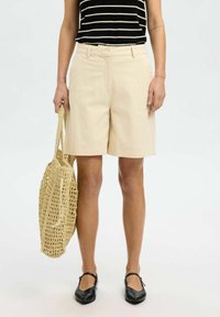 Pantaloni corti in cotone beige con un design sartoriale, dotati di passanti per cintura e tasche frontali. Abbinati a una borsa intrecciata e scarpe nere flessibili.